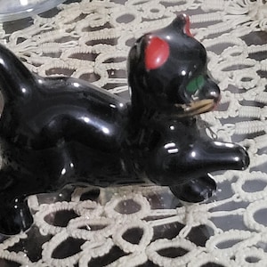 May include: A black ceramic figurine of a cat with red and green painted details. The cat is in a playful pose with its tail raised and its head tilted.
