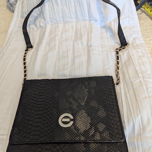 Pode incluir: Uma bolsa de ombro preta com textura de pele de cobra, uma alça de corrente dourada e um logotipo circular prateado com a letra "C" no centro.