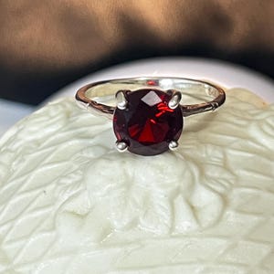 Könnte beinhalten: Ein Silberring mit einem großen, runden, tiefroten Edelstein. Der Edelstein wird von vier Krappen gehalten. Das Ringband hat ein strukturiertes Design. Der Ring wird auf einer weißen, strukturierten Oberfläche präsentiert.