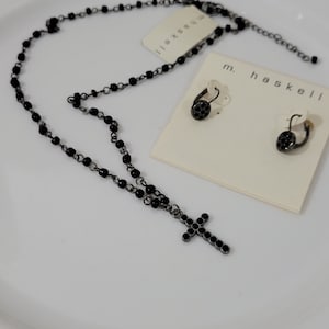 Peut inclure: Un collier noir à perles avec un pendentif en forme de croix et des boucles d'oreilles créoles assorties. Les boucles d'oreilles et le collier sont présentés sur une surface blanche. L'étiquette de la marque indique "m. haskell".