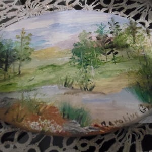 May include: A hand-painted oval-shaped canvas with a landscape scene. The painting depicts a green field with trees and a stream. The sky is blue with white clouds. The painting is signed "Caroline G".