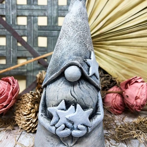 May include: A gray garden gnome statue with a long, pointed hat and a beard. The gnome holds two star-shaped ornaments. The statue has a textured finish and is surrounded by decorative elements, including dried flowers and foliage.
