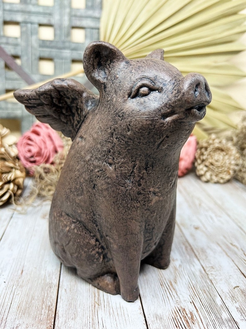 May include: A brown ceramic figurine of a pig with wings. The pig is sitting with its wings spread out and has a cute expression on its face.