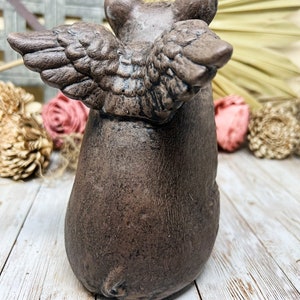 May include: A brown ceramic pig figurine with wings. The pig is standing on a white wooden surface.