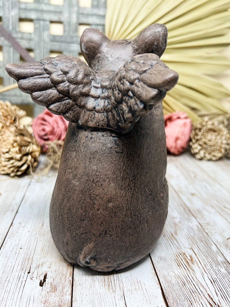 May include: A brown ceramic pig figurine with wings. The pig is standing on a white wooden surface.
