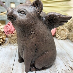 May include: A brown ceramic pig figurine with wings. The pig is sitting on its hind legs with its front legs resting on its belly. The wings are spread out behind the pig.