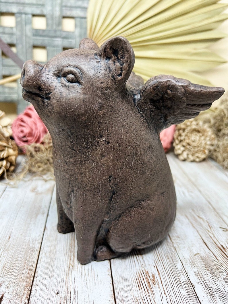 May include: A brown ceramic pig figurine with wings. The pig is sitting on its hind legs with its front legs resting on its belly. The wings are spread out behind the pig.
