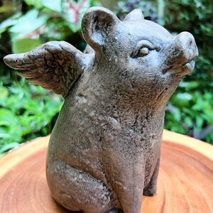 May include: A gray concrete figurine of a pig with wings. The pig is sitting with its head tilted up and its wings spread out behind it.
