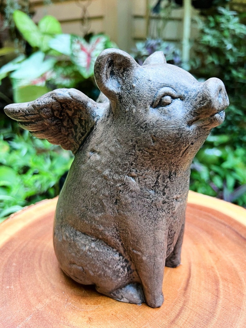 May include: A gray concrete figurine of a pig with wings. The pig is sitting with its head tilted up and its wings spread out behind it.