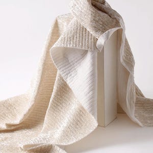 May include: Cream-coloured fabric with a textured, quilted pattern, draped over a white rectangular form. The fabric's edges reveal a contrasting white, ribbed texture. The overall effect is soft and luxurious, suggesting a high-quality textile.