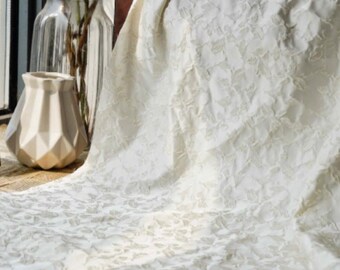 May include: A cream-colored, textured fabric with a floral pattern is draped over a surface. The fabric is soft and flowing, with a subtle sheen. In the background, there are vases with flowers.
