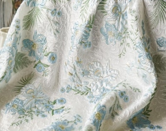 May include: White floral fabric with a raised pattern. The design features light blue flowers and green leaves. The fabric is draped, showing the texture and pattern. The overall look is elegant and suitable for home decor.