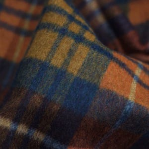 Op de afbeelding: Close-up van een gevouwen stof met een ruitpatroon. De stof heeft een mix van kleuren, waaronder tinten oranje, blauw en bruin. De textuur lijkt zacht, mogelijk wol, met zichtbare geweven details. Het ruitpatroon is een klassiek ontwerp.