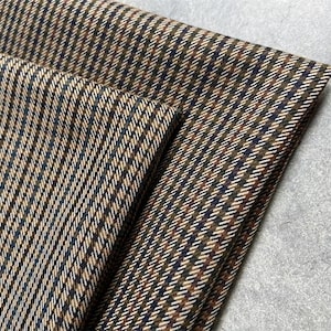 Puede incluir: Primer plano de tela doblada con un patrón de espiga marrón, azul marino y blanco. La tela está tejida apretadamente, con un diseño diagonal. La textura parece suave y adecuada para proyectos de ropa o decoración del hogar.