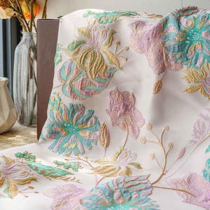 Peut inclure: Tissu crème avec un motif floral dans les tons de turquoise, lavande et or. Le motif présente de grandes fleurs et feuilles stylisées avec des détails complexes. Le tissu semble être un textile décoratif.