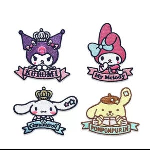 Peut inclure: Quatre écussons brodés représentant des personnages de dessins animés. Les personnages comprennent un chat violet avec une couronne et le nom "Kuromi", un personnage rose avec un nœud et le nom "My Melody", un chien blanc avec une couronne et le nom "Cinnamoroll", et un chien jaune avec le nom "Pompompurin".