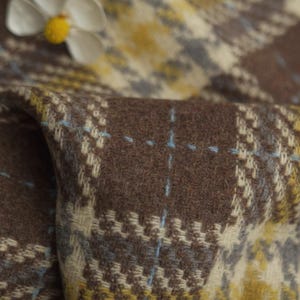 May include: Close-up of a brown, yellow, and blue plaid fabric. The fabric has a textured appearance with a repeating pattern of squares and lines. The colors are muted and earthy, with a soft, warm feel. A white flower with a yellow center is in the upper left corner.