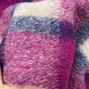 May include: Close-up of a textured fabric with a plaid pattern. The fabric features a mix of colors, including shades of pink, purple, blue, and cream. The material appears soft and possibly woolen, with a slightly fuzzy texture.