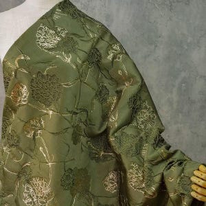 Puede incluir: Tela verde oliva con un estampado floral dorado. La tela está drapeada, mostrando el diseño detallado y la textura. Los elementos florales son intrincados, añadiendo elegancia. Adecuado para diversos proyectos.