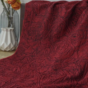May include: A rich crimson fabric featuring a rose pattern. The material is draped, highlighting the texture and design. The roses are outlined in a darker hue, creating a contrast. The fabric is likely for home decor.