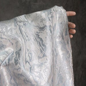 Puede incluir: Primer plano de una tela con un patrón veteado en tonos grises y azul claro. La tela se sostiene, revelando su textura y la forma en que la luz juega sobre su superficie. Los bordes de la tela están sin terminar.