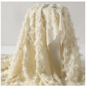 Peut inclure: Tissu crème avec un motif texturé et frangé. Le tissu est drapé sur une forme, mettant en valeur ses détails complexes et sa texture douce. Le tissu semble être un textile décoratif, peut-être pour l'habillement ou la décoration intérieure.