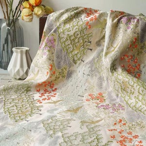 Op de afbeelding: Crèmekleurige stof met een gedetailleerd patroon van bomen, vogels en bloemelementen in groen, oranje en paars. De stof is gedrapeerd, wat het ingewikkelde ontwerp en de textuur benadrukt. Het thema is natuur en landschap.