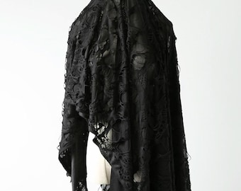 May include: A black, lace-like shawl draped over a form. The fabric has a distressed, torn appearance, creating a unique texture. The shawl is likely part of a gothic or alternative fashion ensemble.