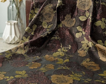May include: A dark fabric with a floral rose pattern in shades of brown, gold, and green. The fabric has a textured appearance and is draped over a surface. The roses are large and detailed, with green leaves.