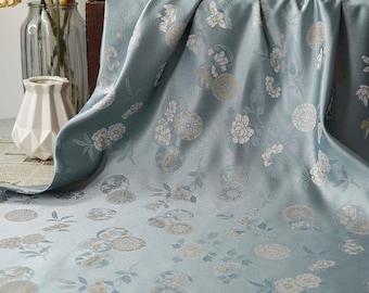 May include: A draped, light blue fabric with a floral pattern. The fabric has a silky sheen and is adorned with white and gold-colored flower and circular designs. The folds of the fabric create shadows, enhancing the texture and visual interest. The overall theme is elegant and decorative.