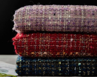 May include: Three rolls of textured fabric in purple, red, and blue. The woven material has flecks of metallic thread. The fabrics are stacked, showing the edges of the rolls. The colours are rich and the texture is detailed.
