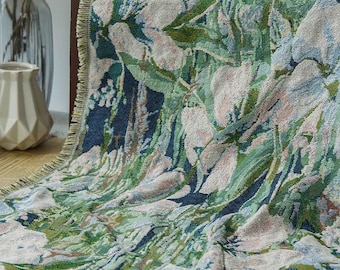 May include: A woven throw blanket with a floral pattern in shades of green, blue, and white. The blanket has a fringed edge and is draped over a piece of furniture. The design features a detailed botanical print, creating a textured and decorative textile.