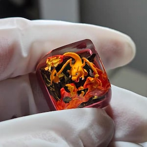 May include: A rectangular, resin keycap featuring a vibrant orange Charizard figure amidst fiery orange and red flames. The keycap has a translucent, pinkish-red border and is set against a white background.