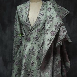 May include: A draped garment on a mannequin, featuring a floral pattern in shades of green and purple. The fabric has a textured appearance, with a deep V-neck and layered design. The overall aesthetic suggests a formal or artistic style.