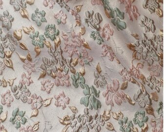 May include: A close-up of a white fabric with a floral pattern. The flowers are pink, green, and gold. The fabric has a textured, raised surface.