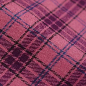 May include: Close-up of a pink plaid fabric with a diagonal pattern. The plaid design features intersecting lines in shades of pink, purple, black, and blue. The fabric has a textured appearance, suggesting a woven material.