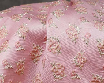 May include: Close-up of a pink fabric with a floral pattern. The fabric has a textured appearance with small, repeating floral designs in a lighter pink and silver. The fabric is folded, creating shadows and depth, highlighting the intricate details of the design.