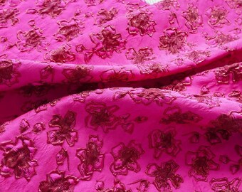 May include: Bright pink fabric with a floral pattern in a darker shade of pink. The fabric is textured and appears to be draped, with visible folds and shadows. The floral design is evenly distributed across the material.