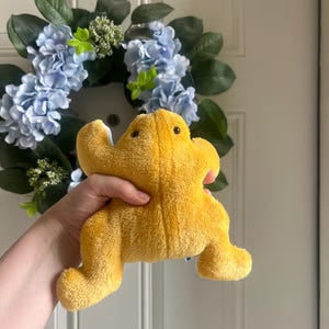 May include: A bright yellow plush frog toy with a soft, textured fabric surface, small dark eyes, and a wide, flat body shape. The stuffed animal features four distinct limbs and a rounded head, showcasing a vibrant golden-yellow color throughout.