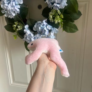 May include: A soft, light pink plush toy shaped like a stylized creature with a smooth, rounded body, two long limbs, and a small dark eye. The plush features a small blue and white tag attached to its side, showcasing a simple, minimalist design.