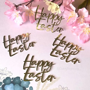 May include: Four gold glitter Happy Easter cutouts on a pink background with pink flowers. The cutouts are in different sizes and styles.