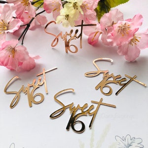 Puede incluir: Confetti de purpurina dorada con las palabras "Sweet 16" en escritura cursiva. El confeti est&aacute; esparcido sobre una superficie blanca con flores rosas.