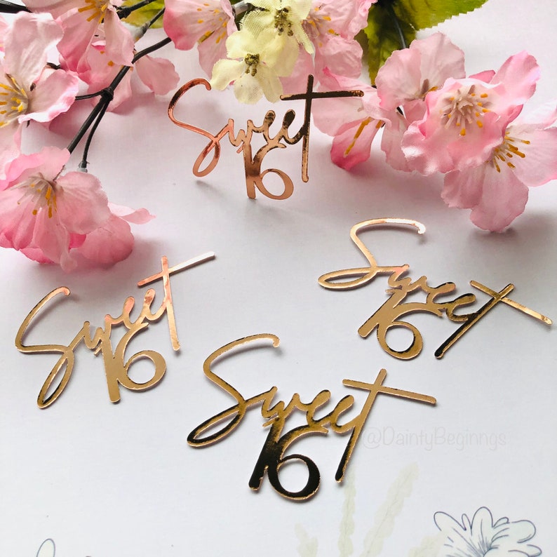 Puede incluir: Confetti de purpurina dorada con las palabras "Sweet 16" en escritura cursiva. El confeti est&aacute; esparcido sobre una superficie blanca con flores rosas.