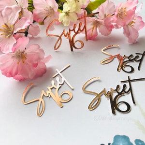 Puede incluir: Cuatro piezas de confeti de oro brillante "Sweet 16" sobre un fondo blanco. El confeti est&aacute; en una fuente de escritura. El confeti est&aacute; rodeado de flores rosas.