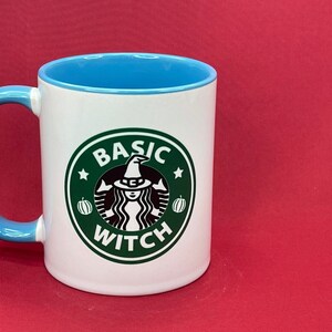 May include: A white ceramic mug with a light blue interior and handle. The front features a circular green logo with a white graphic of a figure wearing a witch hat, flanked by two small pumpkins, with the text BASIC WITCH printed in white.