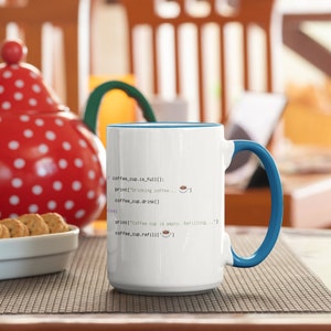Op de afbeelding: Een witte keramische mok met een blauwe binnenkant en handvat, voorzien van zwarte programmeercode tekst die luidt: if coffee_cup.is_full(): print("Drinking coffee... ☕") coffee_cup.drink() else: print("Coffee cup is empty. Refilling...") coffee_cup.refill('☕')