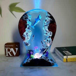 Op de afbeelding: Een blauwe harslamp met een haai, duikers en een gezonken schip erin. De lamp wordt van onderen verlicht, waardoor een blauwe gloed ontstaat. De lamp staat op een tafel met een boek en een houten blok waarop "Resinlamp made by RV" staat.