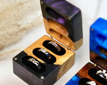 May include: Wooden ring boxes with a purple and black epoxy resin finish, each holding three rings on a black velvet interior. The boxes are open, displaying the rings. The boxes are made of wood and epoxy resin.