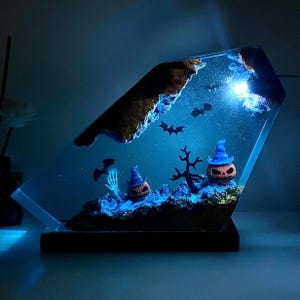 May include: A Halloween resin art piece with two pumpkin lanterns in blue witch hats, a skeleton hand, and bats. The scene is lit with blue light, creating a spooky atmosphere. The art piece is encased in a clear, angled resin block.