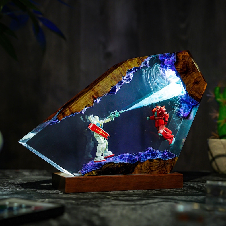 May include: A unique resin art piece featuring two robot figures in a battle scene. The artwork is encased in a clear, angled resin block with a wooden frame. The scene is illuminated with blue and white light effects, creating a dynamic display.